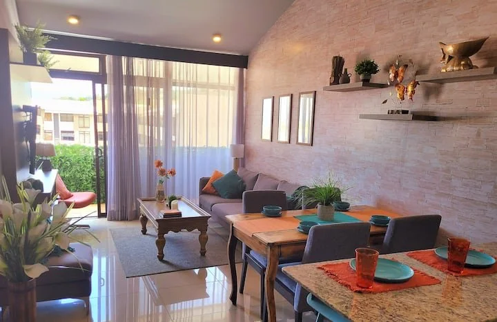 Living room with dining area, gray sofa with colorful pillows, glass sliding door with curtains leading to balcony, coffee table with flowers, decorative shelf with ornaments, and a table set with teal plates and orange glasses.