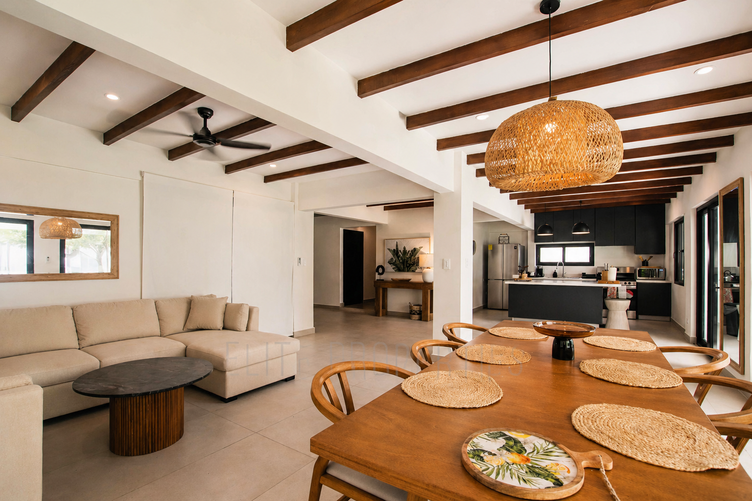 Open-concept living and dining area with white walls, wooden ceiling beams, beige sectional sofa, black coffee table, wooden dining table with woven placemats, hanging rattan pendant light, and a kitchen with black cabinets, stainless steel appliance