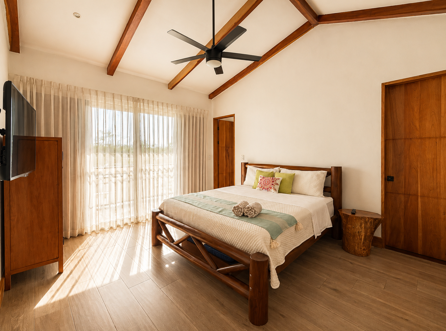 A bedroom with a large wooden bed, beige bedding, and green and patterned pillows. There is a wooden wardrobe, a small side table with a remote, a ceiling fan, and large window with sheer curtains. The room has wooden beams and light-colored tile flo