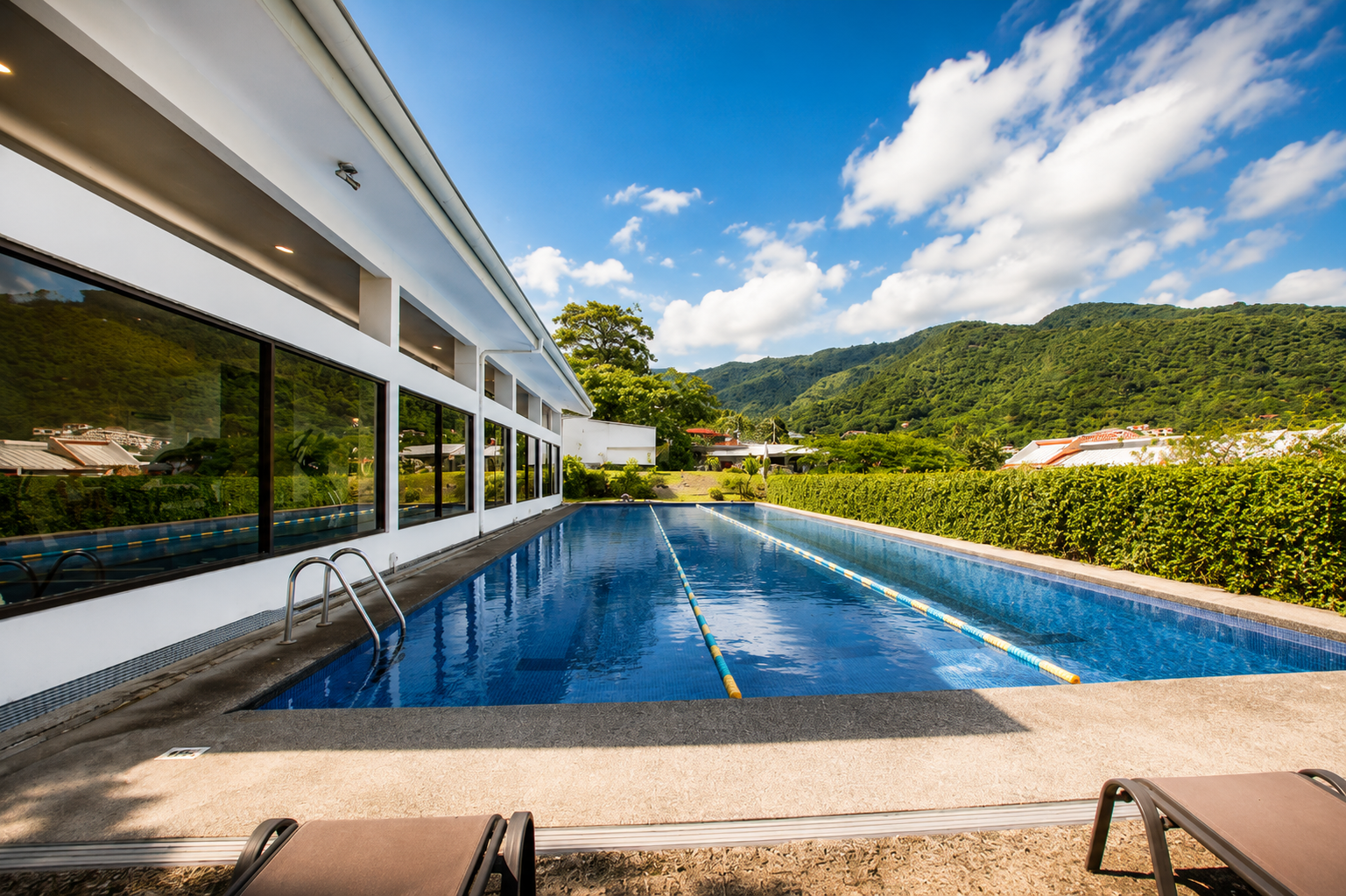 An outdoor swimming pool next to a modern white building with large windows, surrounded by greenery and mountains in the background, under a blue sky with clouds.