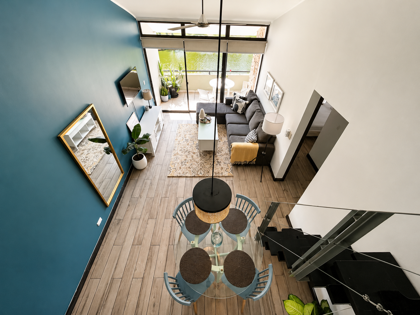 A living room with a blue accent wall, a white TV stand, and a gray sectional sofa facing a glass door balcony overlooking a body of water. There is a white coffee table, a patterned rug, and framed art on the walls. A yellow blanket and pillows are 