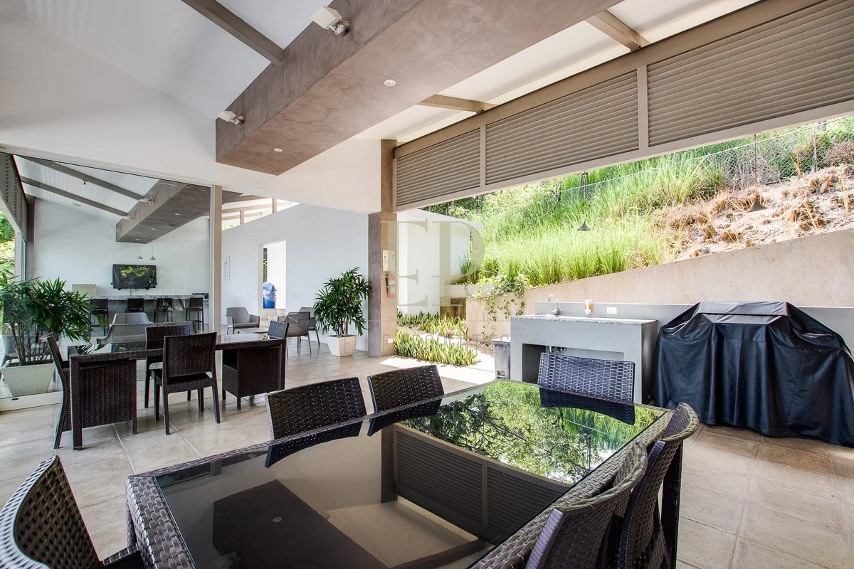 Indoor-outdoor dining area with glass-top table, wicker chairs, potted plants, and a covered barbecue grill, with a view of a garden on a sunny day.