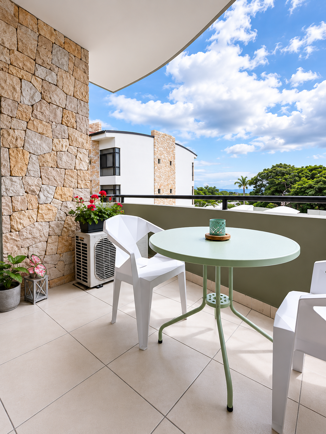 Balcony with beige tiled floor, white plastic chairs, a green round table, potted plants, a flower pot with pink and red flowers, an air conditioning unit, and a view of a modern building, trees, and a partly cloudy sky.