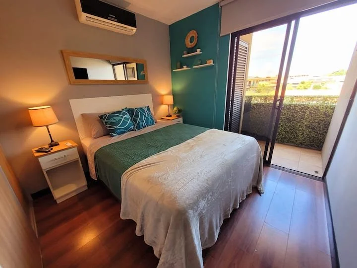 A bedroom with a double bed, two side tables with lamps, a mirror on the wall, and a sliding glass door leading to a balcony with a view of trees and buildings.