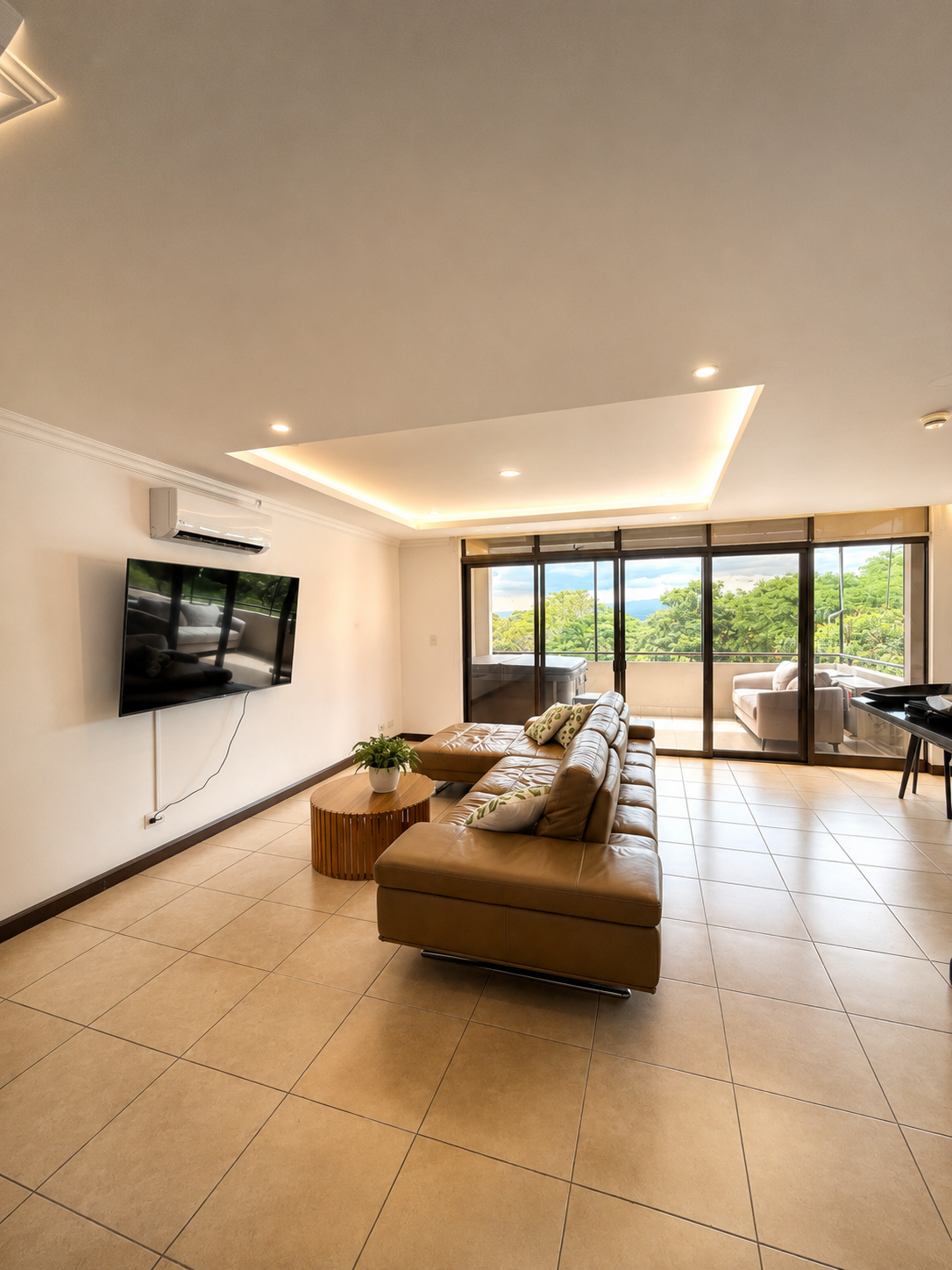 Living room with tan leather sectional sofa, round wooden coffee table with a potted plant, wall-mounted flat-screen TV, sliding glass doors leading to a balcony with outdoor seating, trees and blue sky outside.