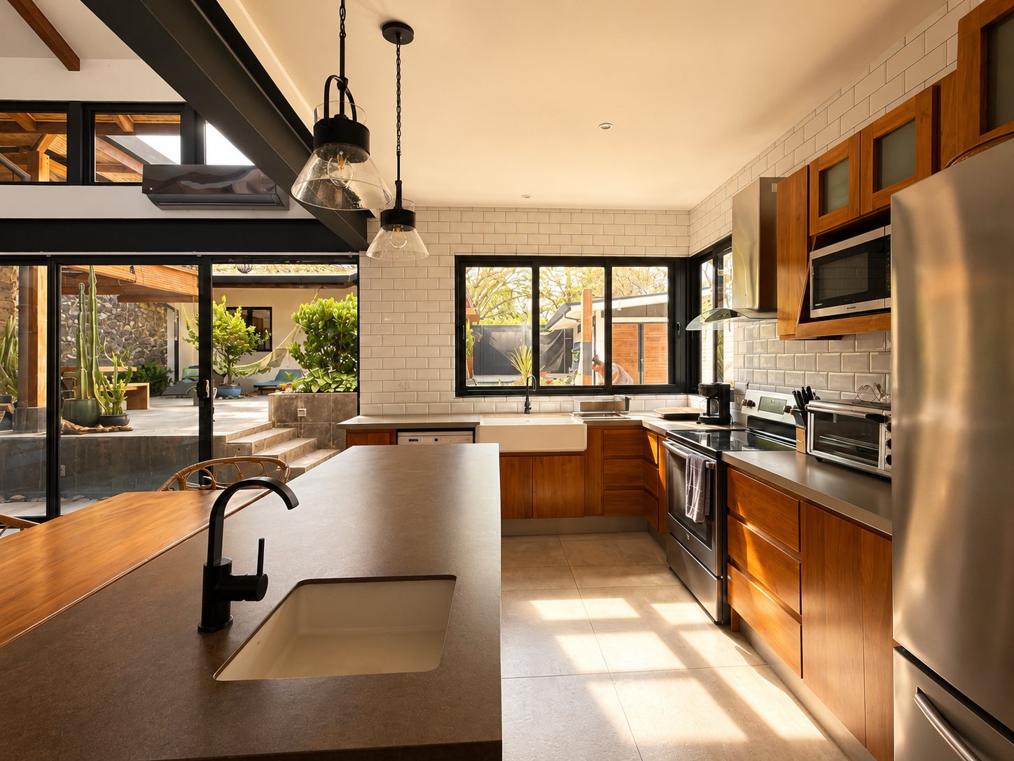 Modern kitchen with white brick walls, wooden cabinets, black appliances, and large windows overlooking an outdoor patio with plants and stone steps.