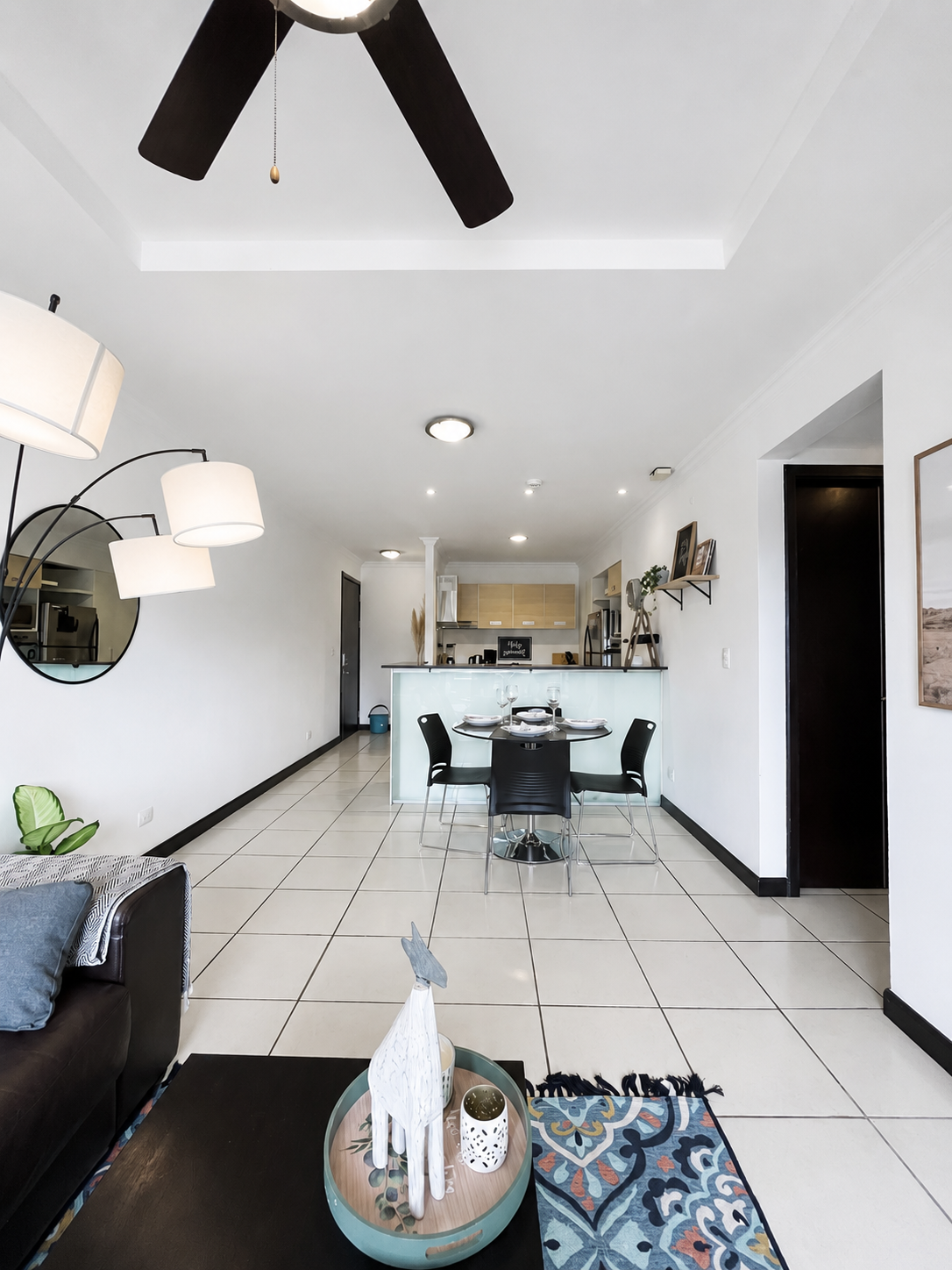 Living room with a black sofa, a wooden table with decorative white animal sculpture, colorful patterned rug, and white tiled floors. Dining area with a round black table and four black chairs, situated in an open space with a kitchen in the backgrou