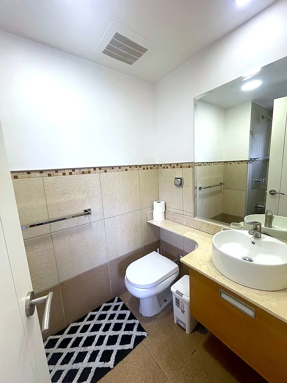 Small bathroom with a white toilet, beige countertop with a round white sink, large mirror, towel bar, and built-in storage with a roll of toilet paper and trash bin.