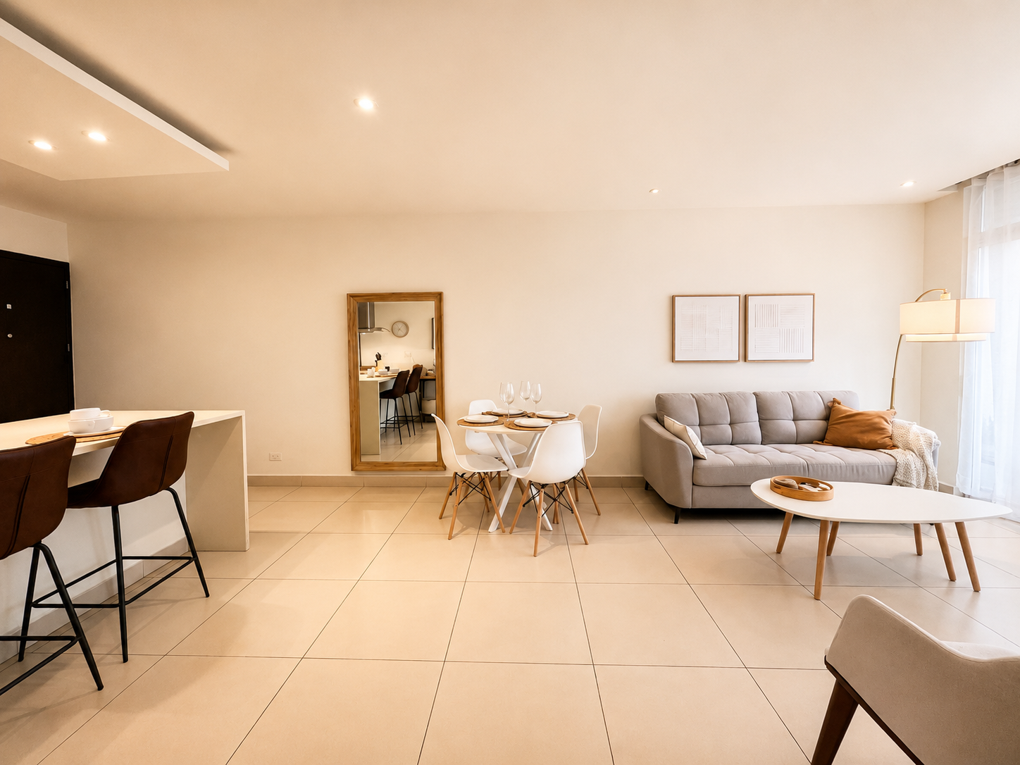 Modern living room with dining area, gray sofa, round dining table with four white chairs, and a large mirror on the wall.