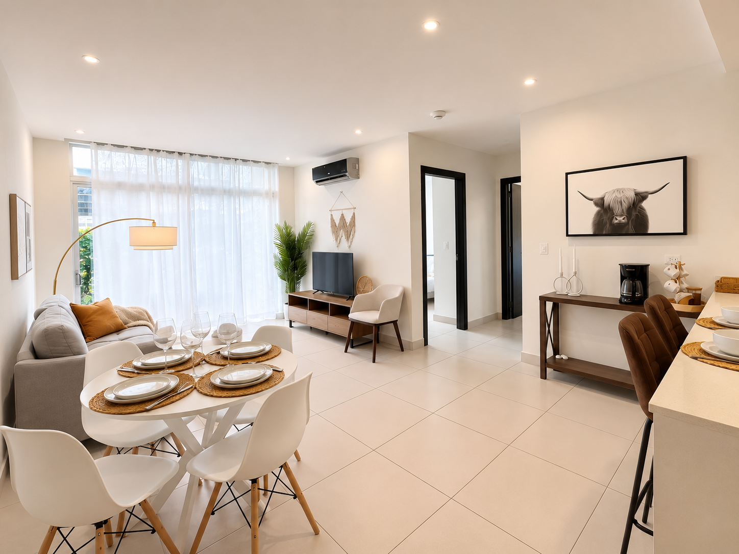 Modern living room with a dining table set for four, a sofa with pillows, a TV on a stand, a white armchair, a large window with sheer curtains, a potted plant, and a black and white highland cow wall art.