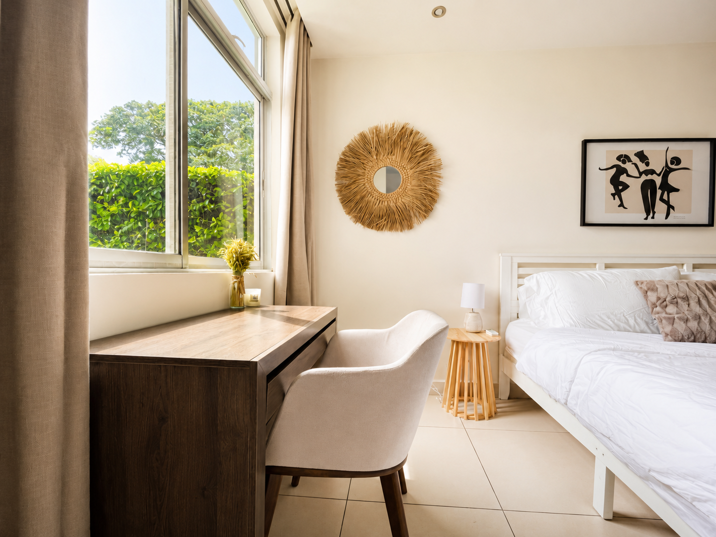 A cozy bedroom with a white bed, a nightstand with a lamp, a wooden desk and chair near a window with green foliage outside, wall art including a round straw mirror and a black and white abstract picture.