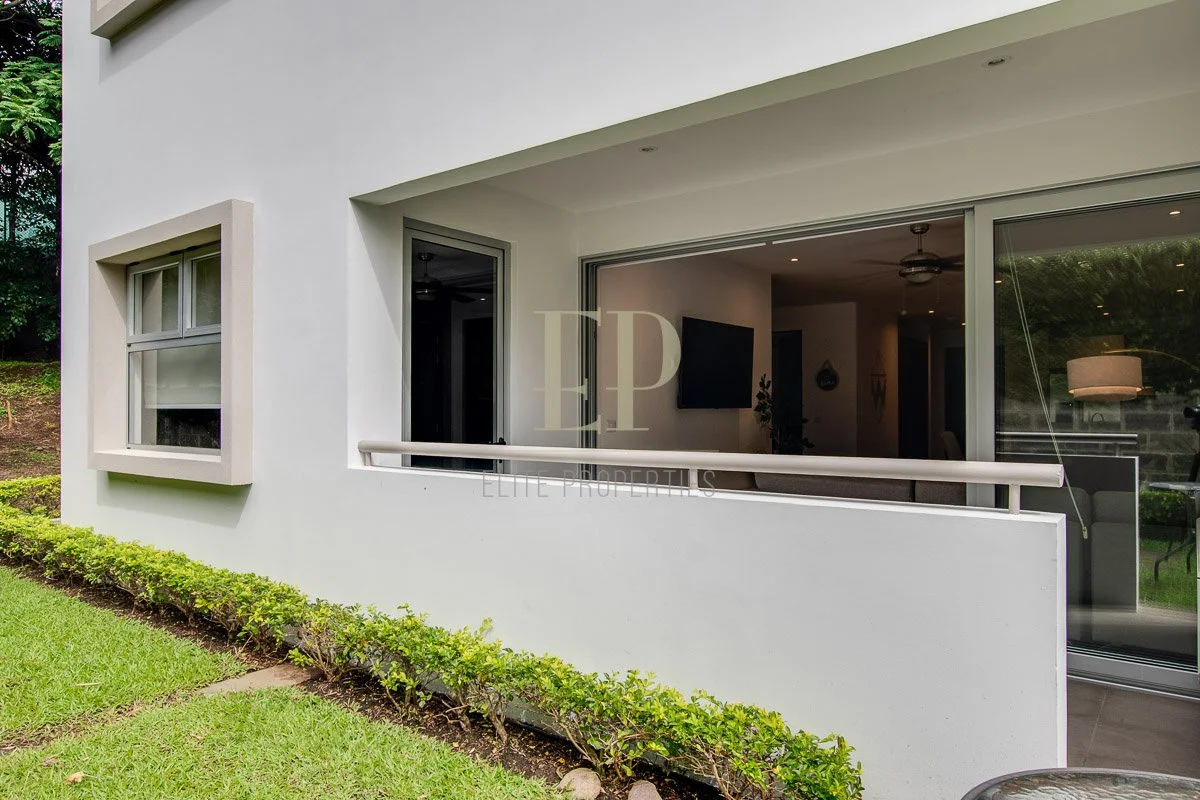 Exterior view of a modern white house with a sliding glass door revealing the interior living room and a window with a blind, surrounded by green grass and bushes.