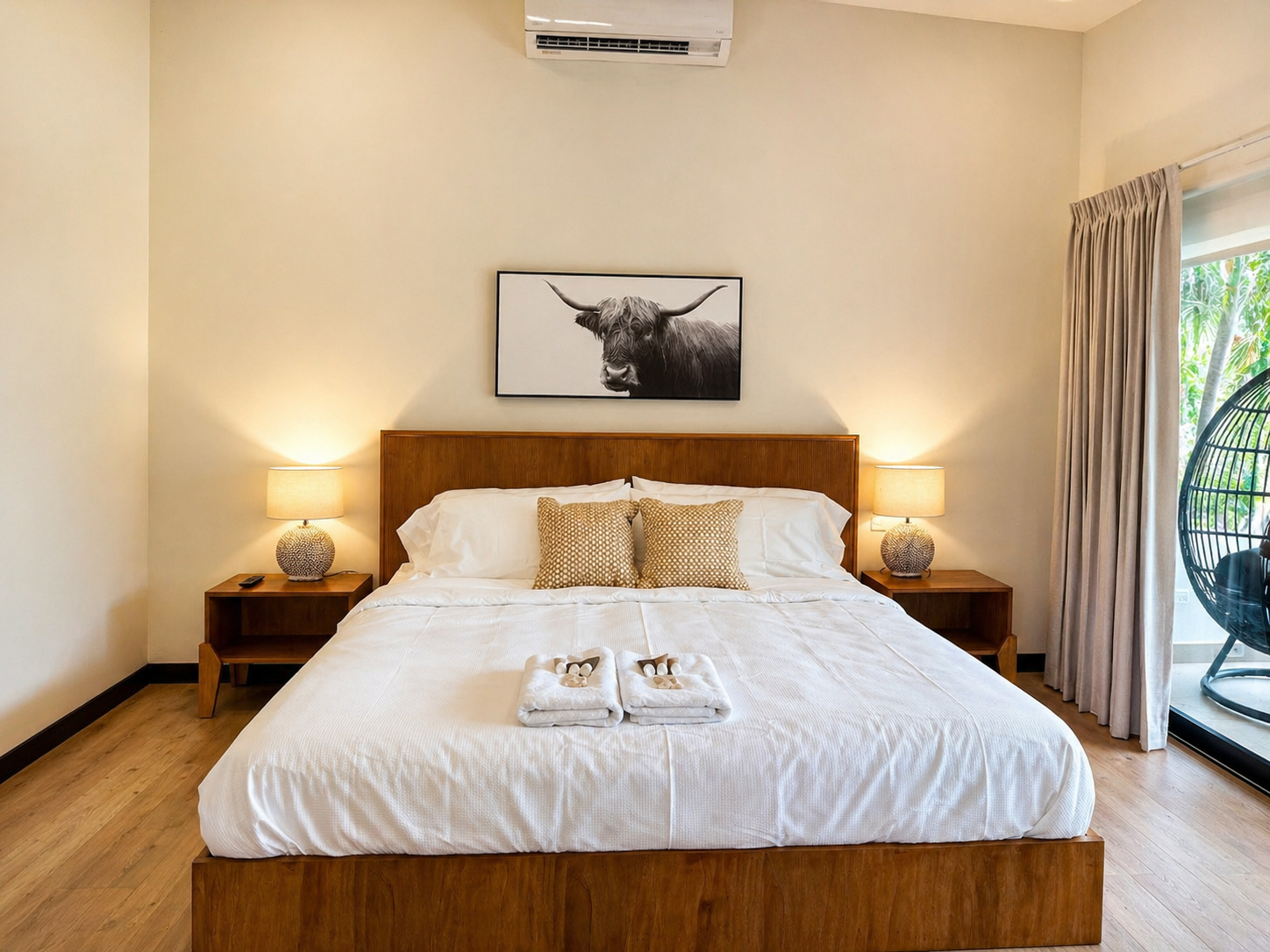 A neatly made bed with white linens and two beige pillows, flanked by two matching bedside tables with lamps, in a bright hotel room with wooden flooring, a black and white bull picture above the headboard, and balcony door with curtains and a hangin