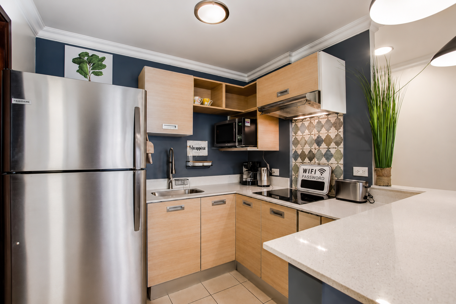 Modern kitchen with stainless steel refrigerator, beige cabinets, white countertops, blue accent wall, microwave, coffee maker, toaster, and a tall green plant in a woven basket.