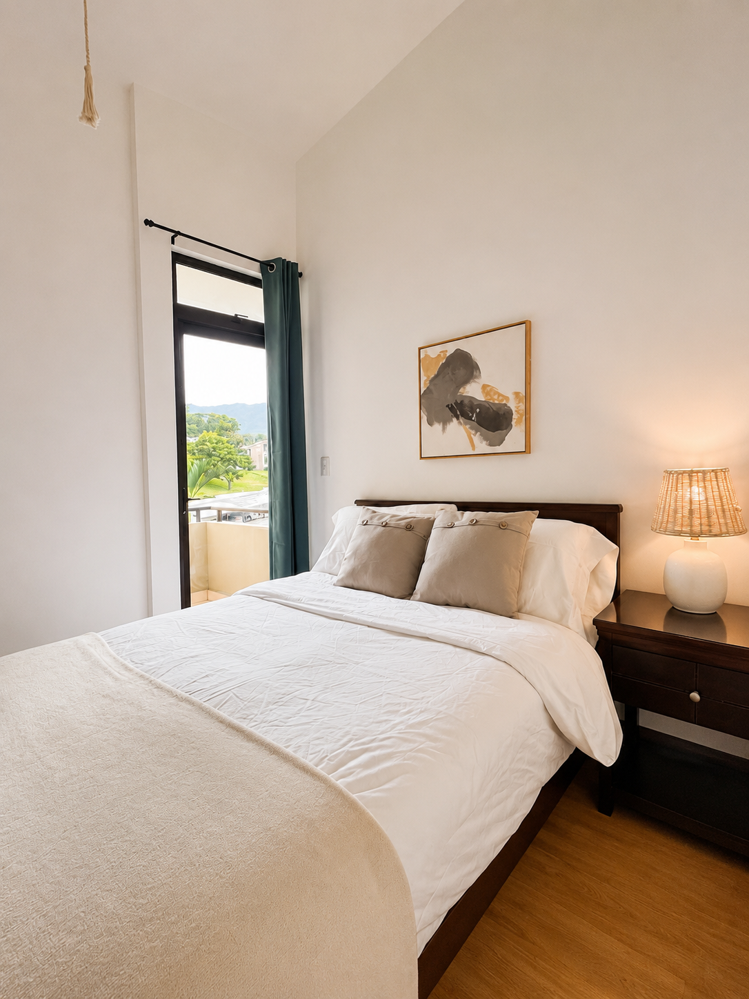 A neatly made bed with white bedding and beige pillows, a dark wooden headboard, and a nightstand with a beige lamp, in a bright room with a sliding glass door leading to a balcony and showing a view of trees and a blue sky outside.
