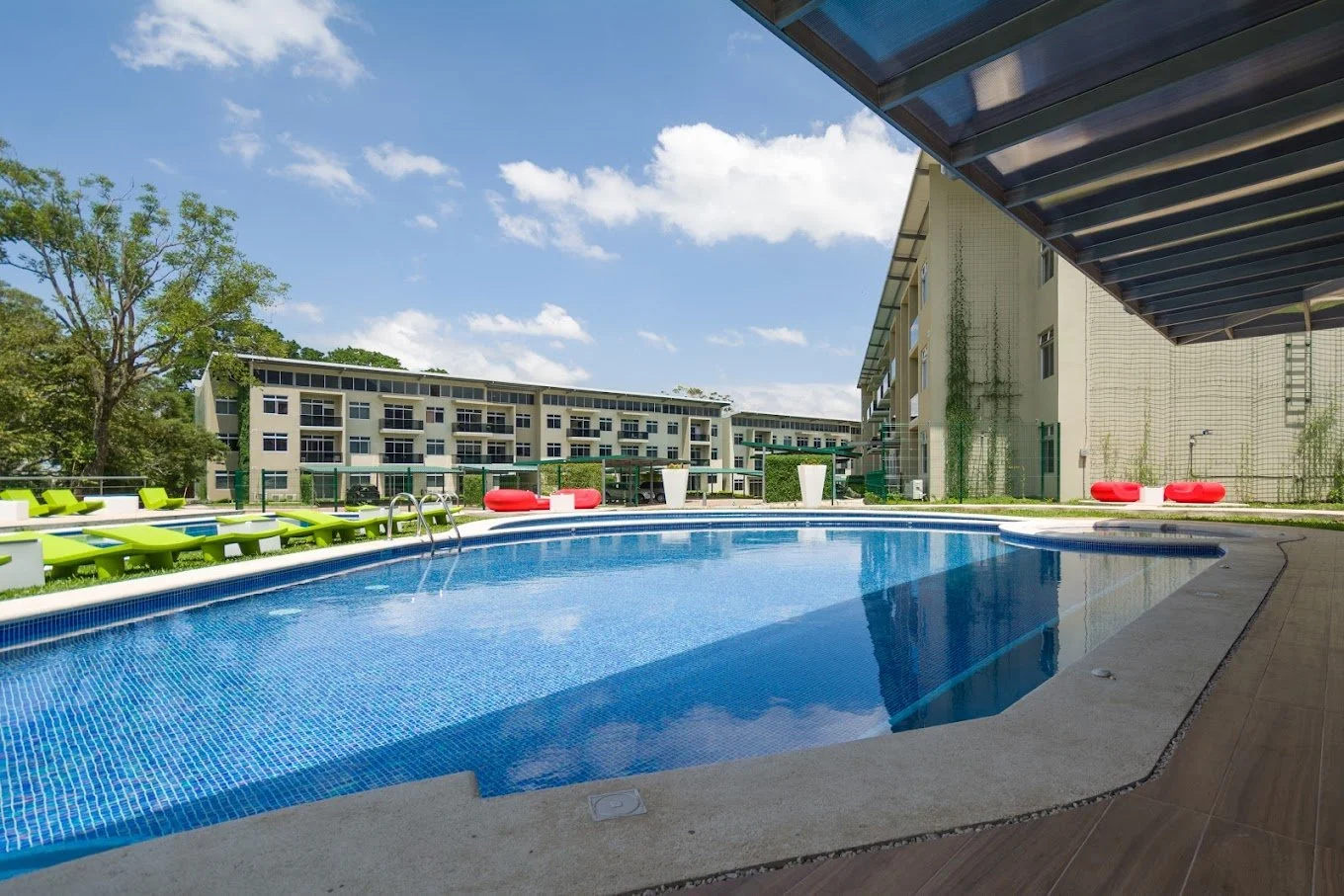 Empty outdoor swimming pool with blue tiles, green lounge chairs, red bean bag chairs, surrounded by residential buildings and trees on a sunny day.
