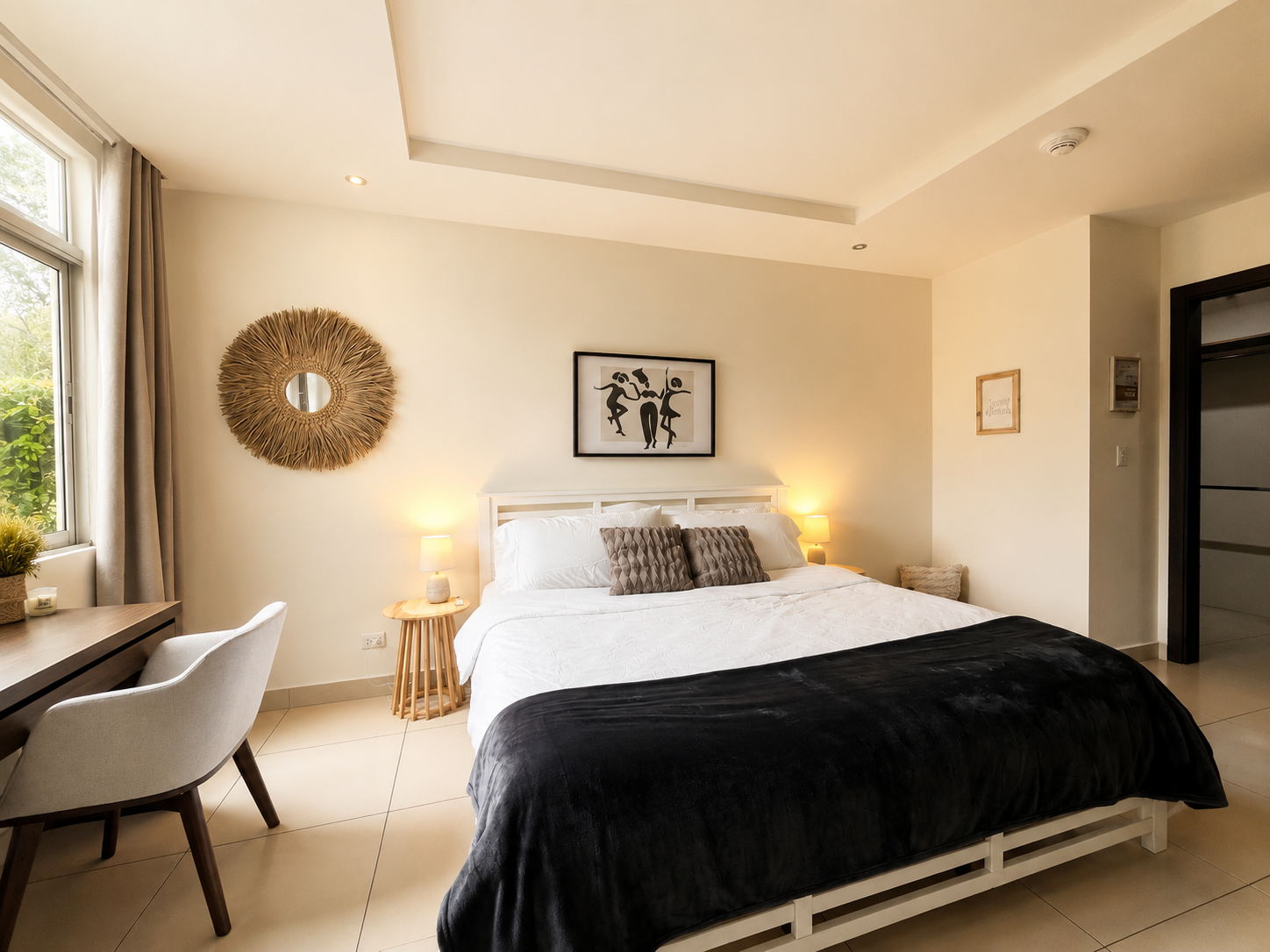 A bedroom with a white bed, beige walls, black and gray pillows, a black blanket, wooden side tables with lamps, a framed artwork above the bed, a round decorative mirror on the wall, a window with blinds and curtains, and a desk with a white chair.