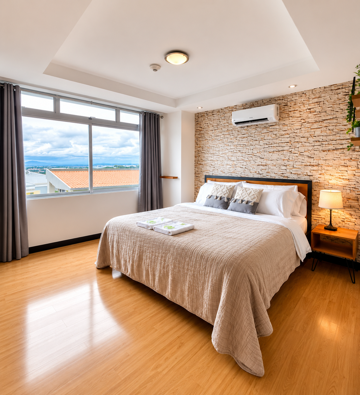 A modern bedroom with a large bed, beige bedding, decorative pillows, a nightstand with a lamp, a brick accent wall, a large window with curtains showing an outdoor view, and an air conditioning unit mounted on the wall.