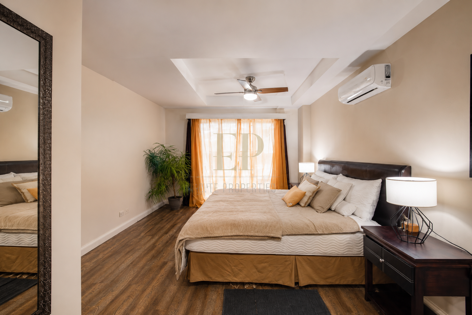 Modern bedroom with a large bed, beige bedding, and multiple pillows. Nightstand with a lamp, a potted plant near the window with orange curtains, hardwood flooring, and a ceiling fan.
