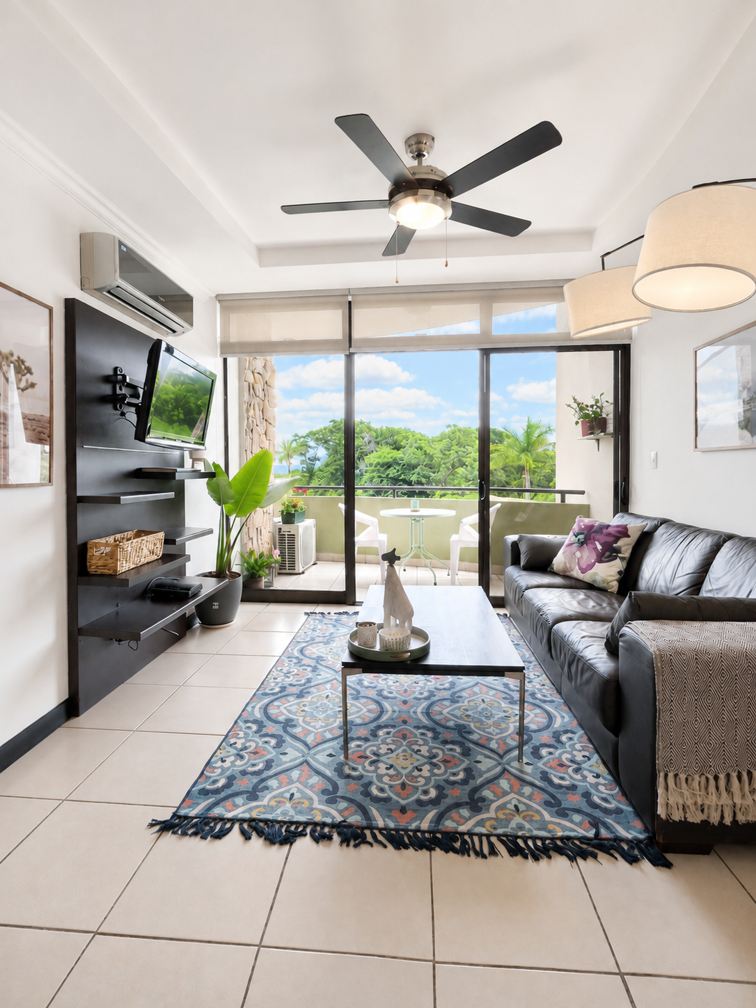 Living room with black leather sofa, glass sliding doors opening to a balcony with outdoor furniture, TV mounted on black wall panel, ceiling fan, potted plants, decorative rug, and ceiling-mounted lamps, overlooking lush greenery.