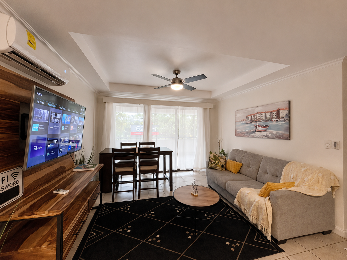 Living room with gray sofa, yellow cushions, rectangular wooden coffee table, black area rug, TV on wooden media console, dining table with four chairs near large window with white curtains, wall art of boats and harbor, ceiling fan, air conditioning