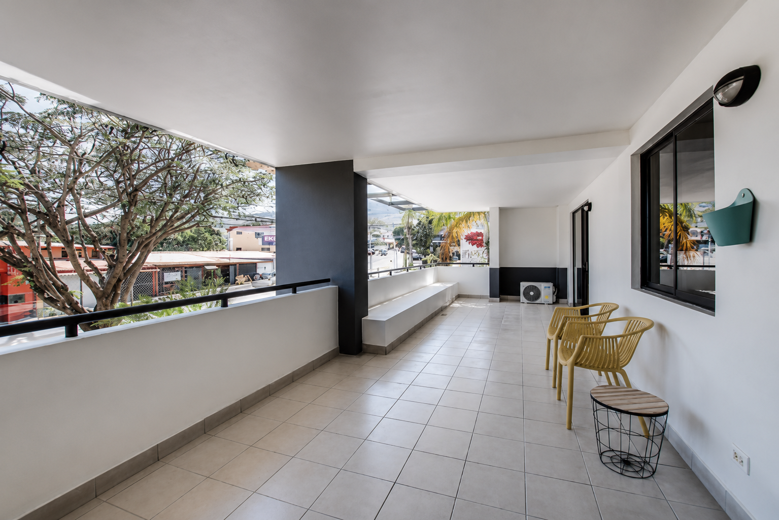 Empty balcony with beige tiled floor, white walls, black window frame, yellow chairs, and a small black wire table, overlooking a street with trees and buildings.