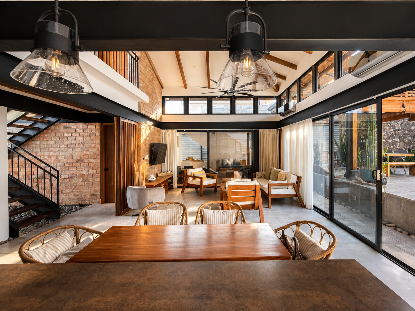 A spacious, modern living room with large glass windows and sliding doors, brick walls, and wooden accents, furnished with sofas, armchairs, and a wooden dining table.
