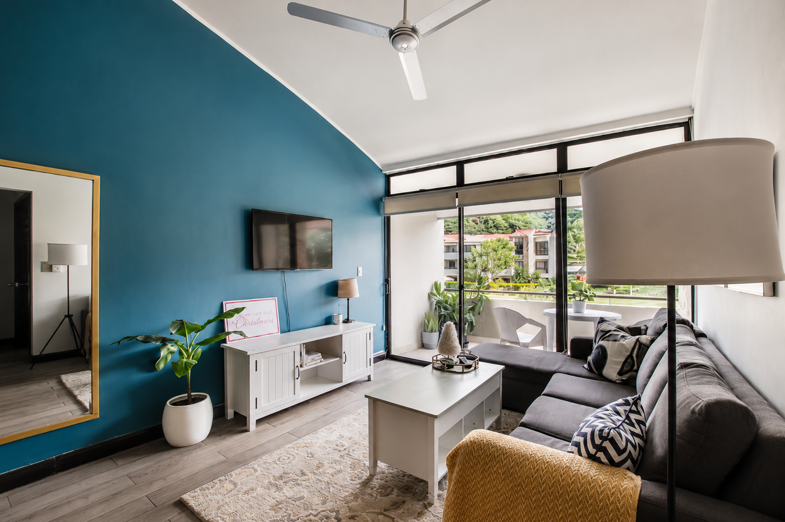 Living room with a blue accent wall, a gray sectional sofa with patterned pillows, a white coffee table, and a large floor lamp. There's a TV mounted on the wall, a white media console, and potted plants near the large glass sliding door leading to a