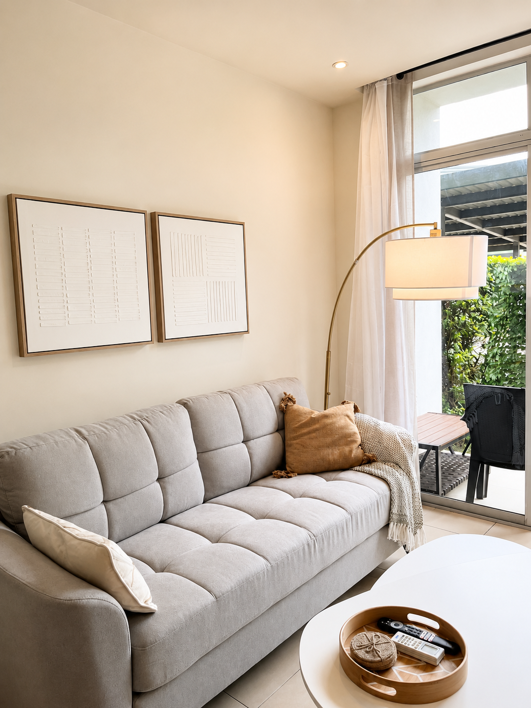 Living room with a beige sofa, two decorative pillows, a white coffee table with remote controls, and wall art with white textured frames. There is a floor lamp with a white shade, and a sliding glass door with curtains leading to a balcony with outd