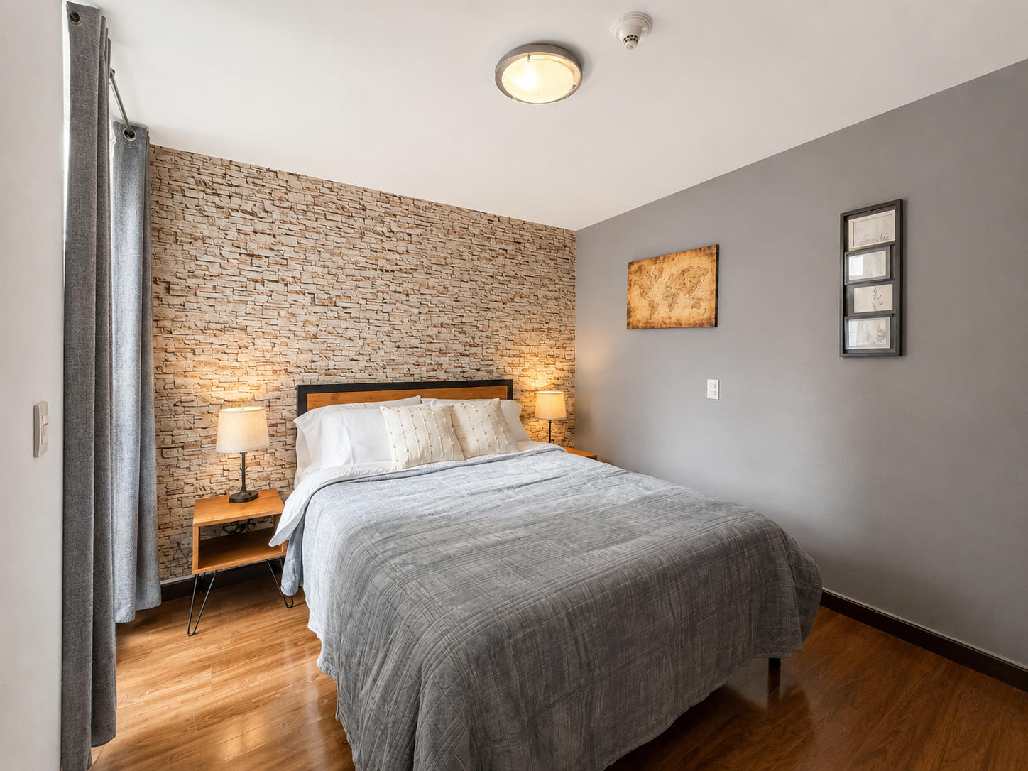 A bedroom with a bed, bedside tables with lamps, a stone accent wall behind the bed, gray walls, framed artwork, hardwood flooring, and gray curtains.