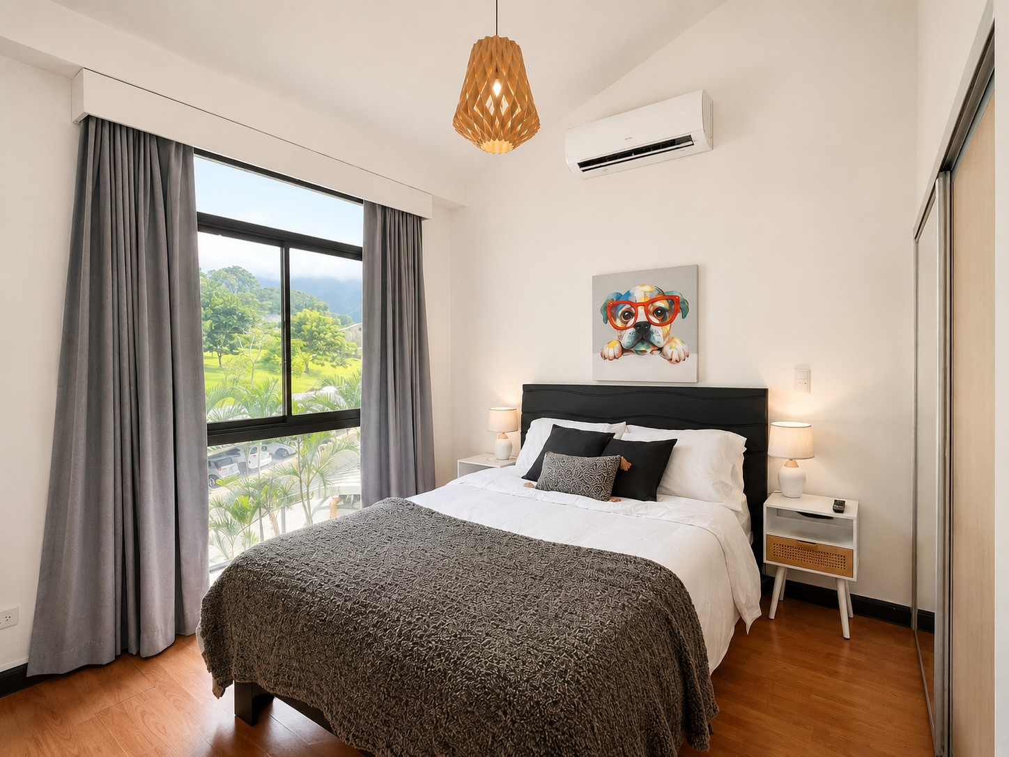 A modern bedroom with a large window showing a scenic view of green trees and mountains, a black bed with white bedding and multiple pillows, framed dog artwork above the bed, two white bedside tables with lamps, an air conditioner, and a wooden clos