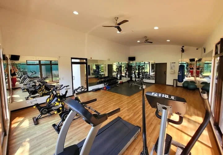 Empty gym room with cardio machines like treadmills and exercise bikes, large mirrors on the walls, and boxing equipment in the background.