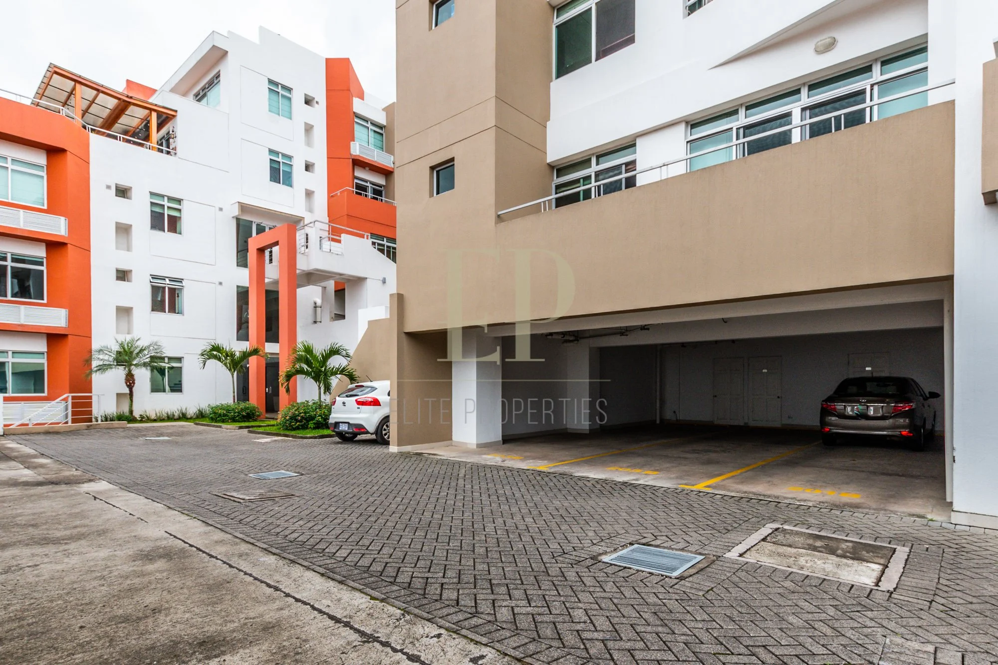 Modern apartment complex with parking area, two parked cars, and palm trees in the background.
