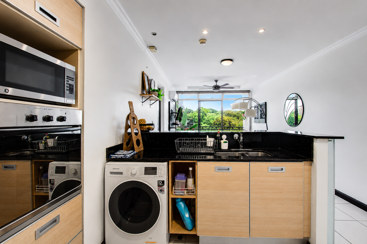 View of a modern kitchen with wood cabinets, black countertops, a washing machine, and a view of greenery outside through large windows.