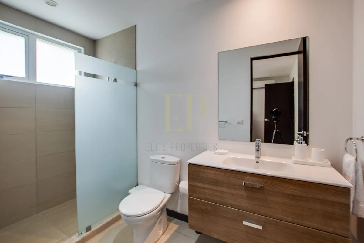 Modern bathroom with a toilet, wooden vanity with a white sink, a large mirror, and a glass shower enclosure, illuminated by natural light from a window.