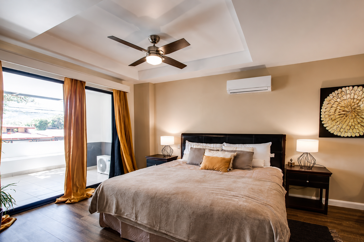 Modern bedroom with large bed, beige bedding, decorative pillows, bedside tables with lamps, sliding glass doors with gold and black curtains leading to balcony, wall-mounted air conditioning unit, ceiling fan, artwork on wall, and natural light comi