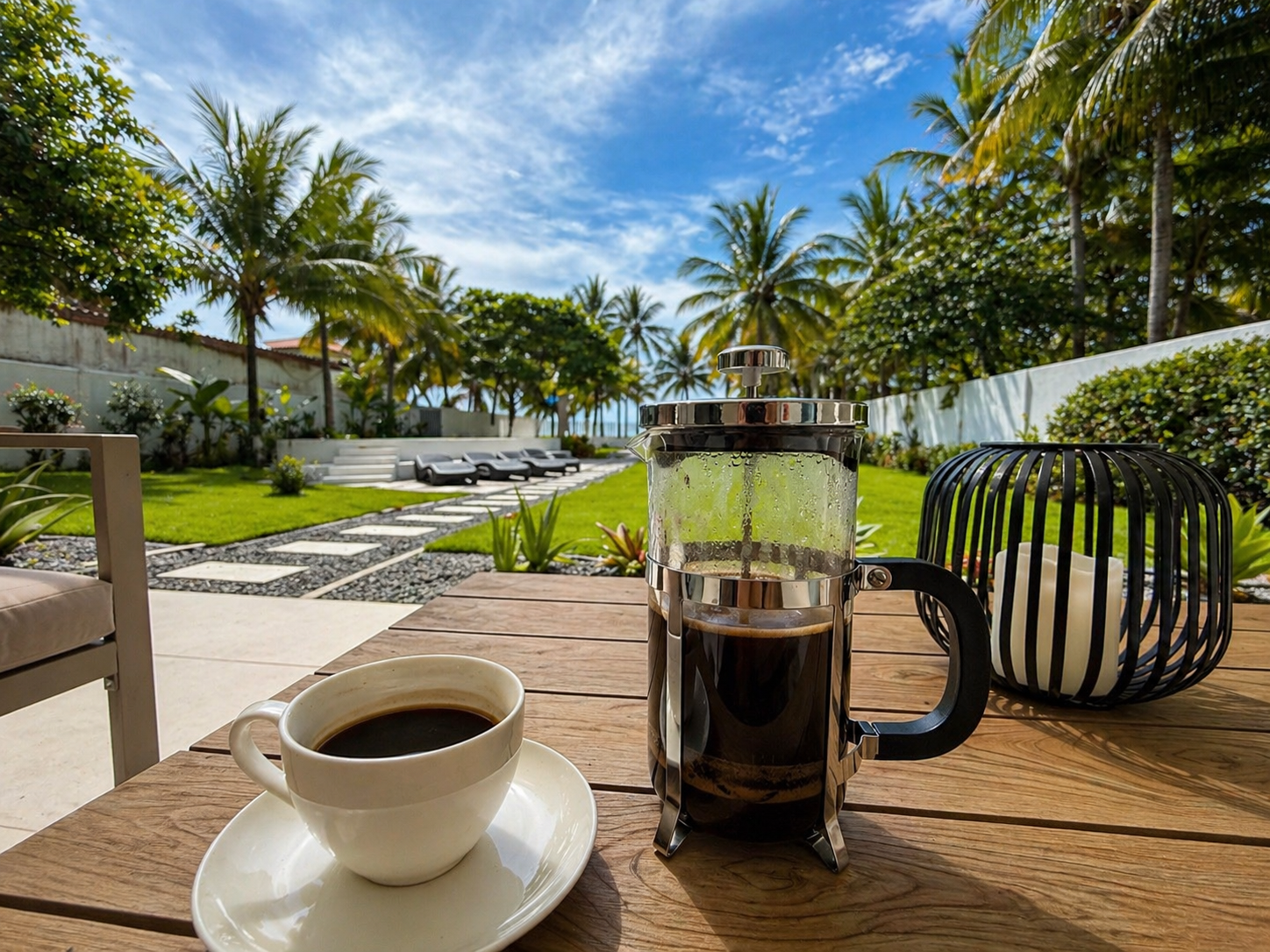 Outdoor patio scene with a French press coffee maker, a cup of black coffee on a saucer, and a decorative lantern on a wooden table, with a lush garden, palm trees, sun loungers, and a blue sky in the background.