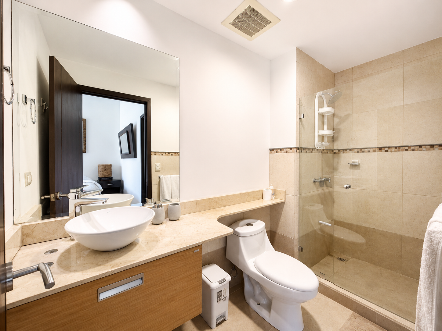 A modern bathroom with a large mirror, vessel sink on a marble countertop, toilet, and a walk-in shower with beige tiles and a glass partition.