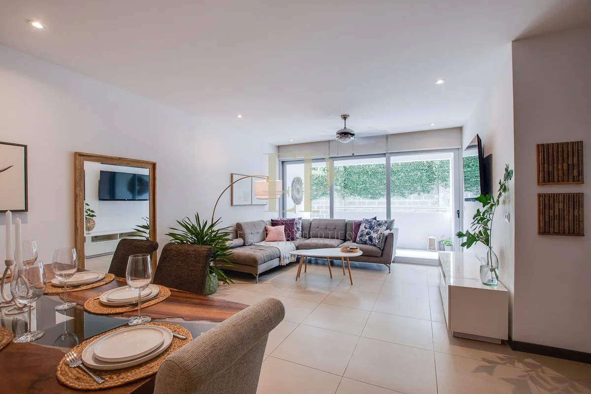 Modern living room with a gray sectional sofa, a small wooden coffee table, a large window with blinds, a wall-mounted flat-screen TV, and decorative pillows, with a dining table set for four in the foreground.