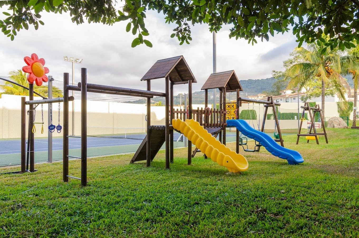 A colorful playground with slides, swings, and climbing structures on a grassy field, with palm trees and a tennis court in the background.