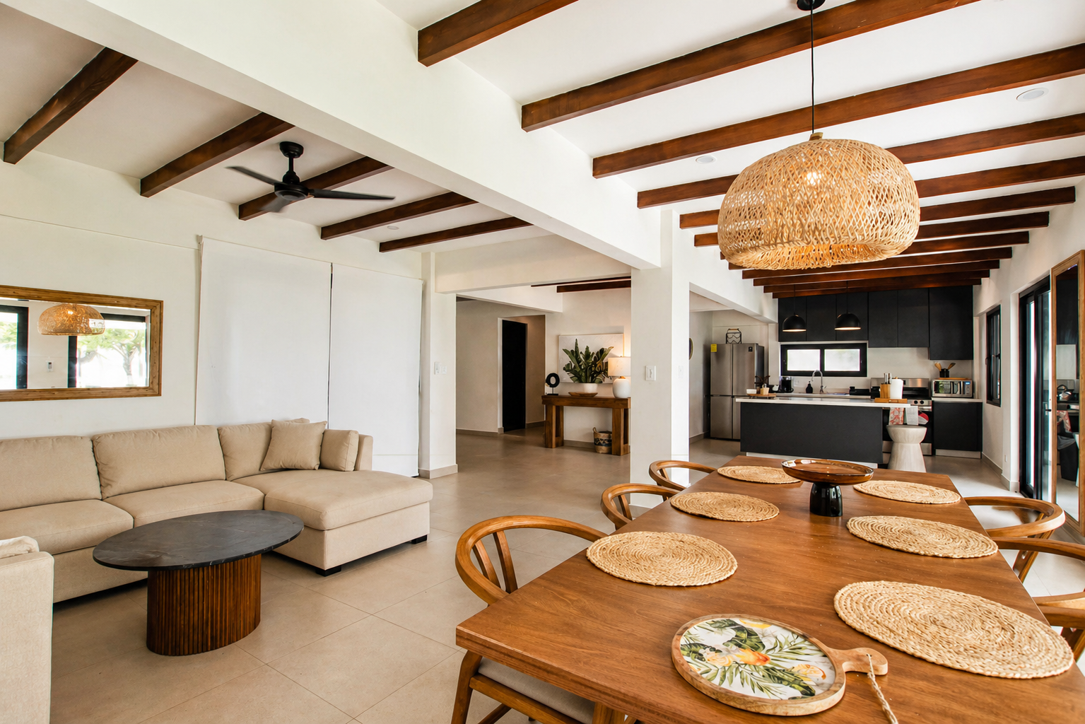 Open-concept living and dining area with beige sectional sofa, wooden dining table with woven placemats, and a modern kitchen with dark cabinets and stainless steel appliances. Exposed wooden ceiling beams and wicker pendant light fixtures.
