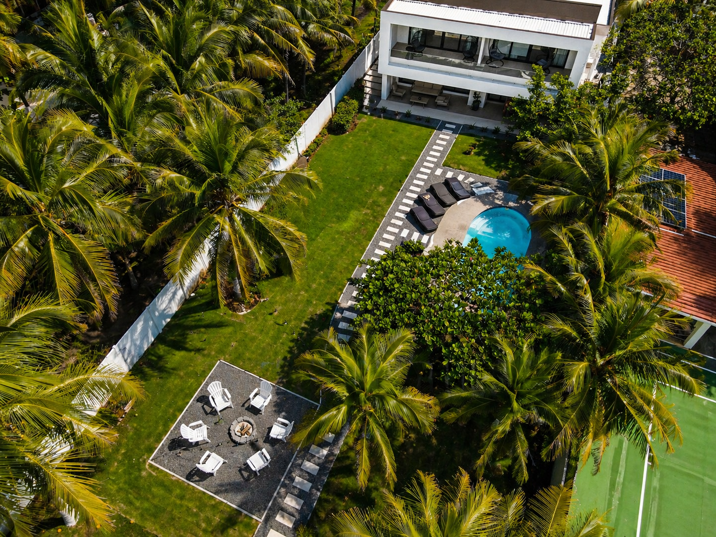 Aerial view of a backyard with tall palm trees, a grassy lawn, a small swimming pool, outdoor seating area with white chairs and fire pit, and a modern white multi-story house.