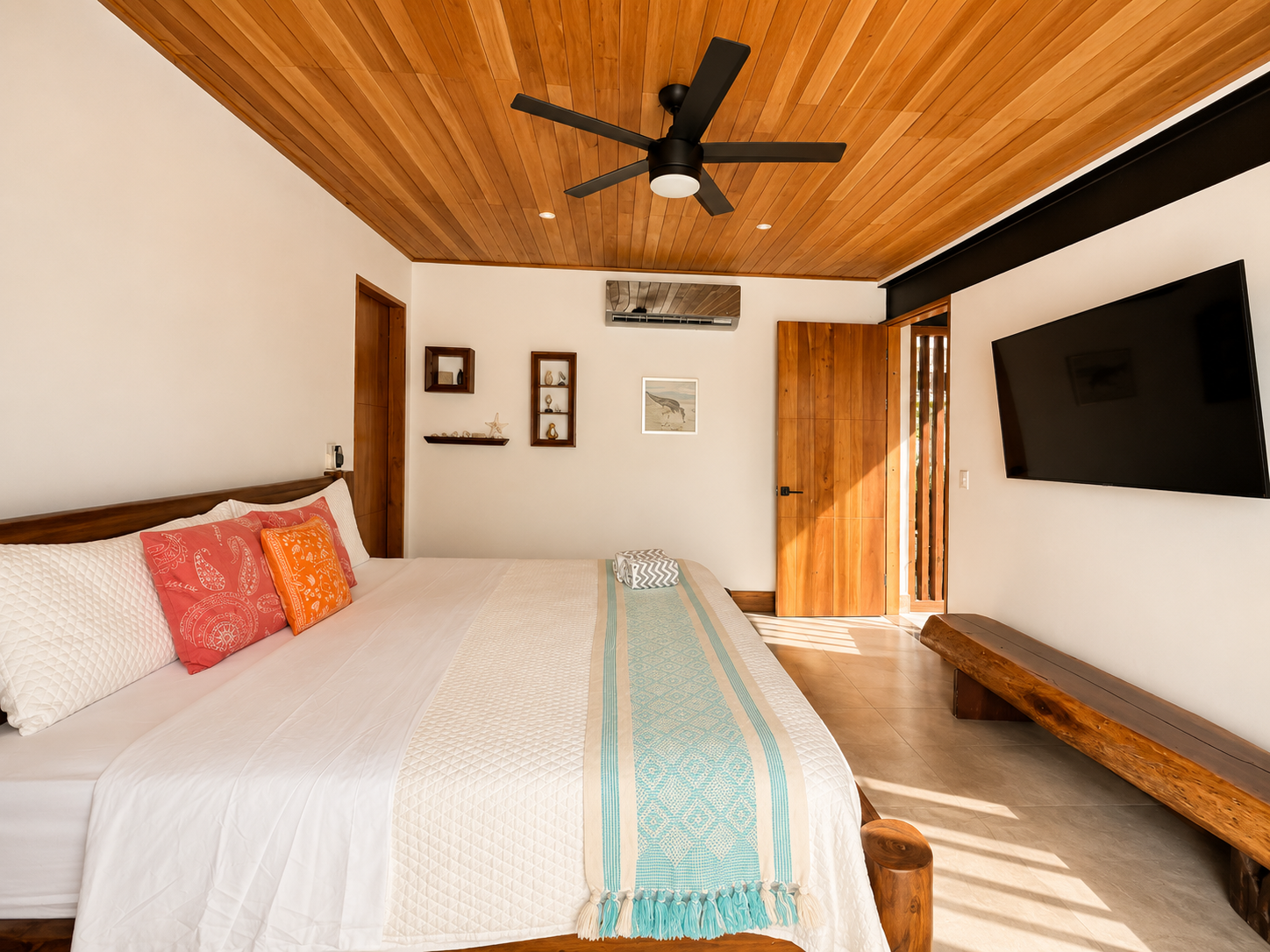 A bedroom with a wooden ceiling, a large bed with white and orange pillows, a wall-mounted TV, and a wooden bench near the door.