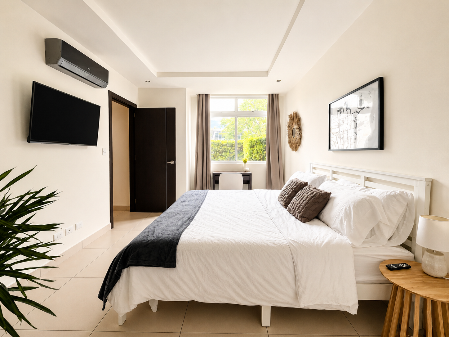 Bright, modern bedroom with a large bed, white bedding and throw pillows, a window with curtains, a small desk and chair, wall-mounted TV, air conditioner, artwork, and a bedside table with a lamp.