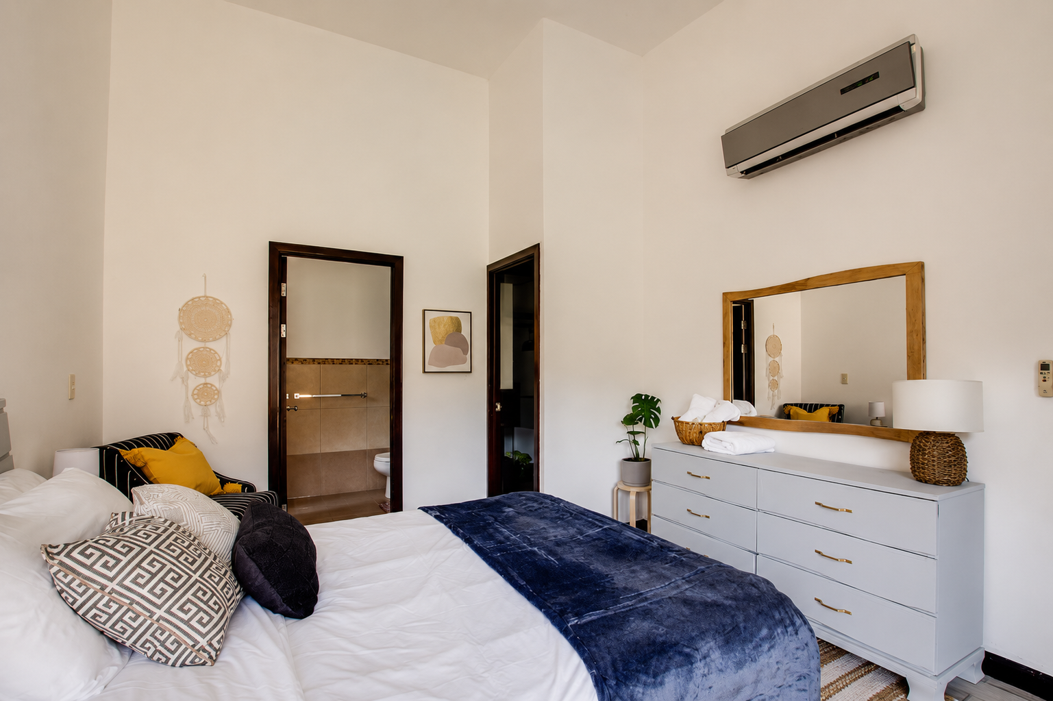 A bedroom with a bed, a dresser with a mirror, a plant, and an air conditioning unit on the wall.