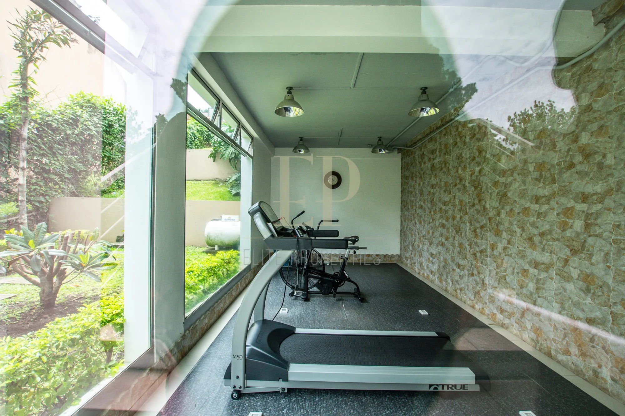 Treadmill in a gym room with large windows overlooking a green garden and stone wall interior.