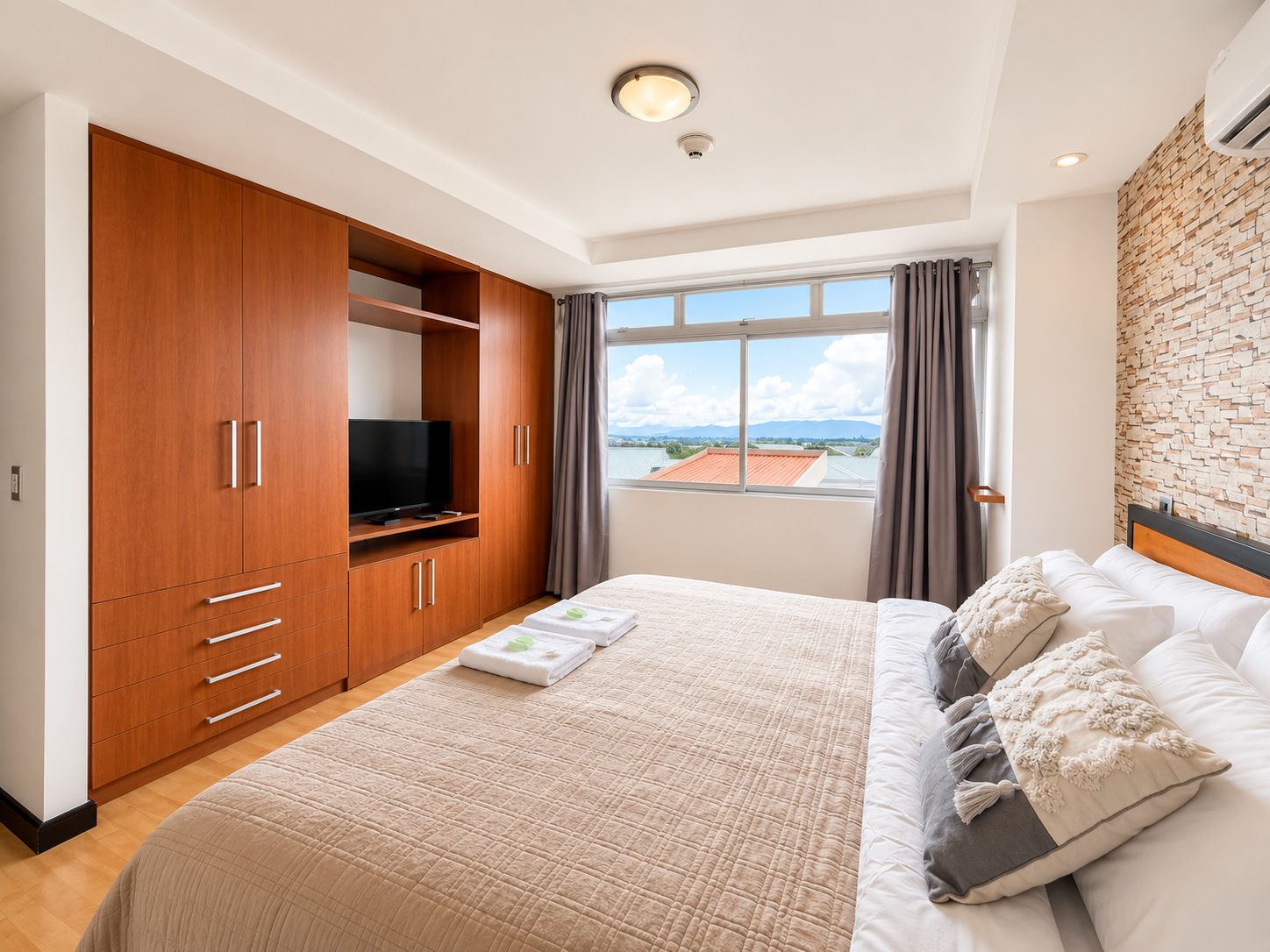 Hotel bedroom with a large bed, pillows, wooden wardrobe, TV, and a window with a view of rooftops and mountains.
