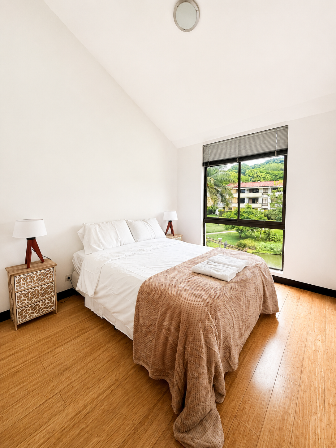 A modern bedroom with a white bed, two bedside tables with lamps, large window revealing greenery and a building outside, and wooden flooring.