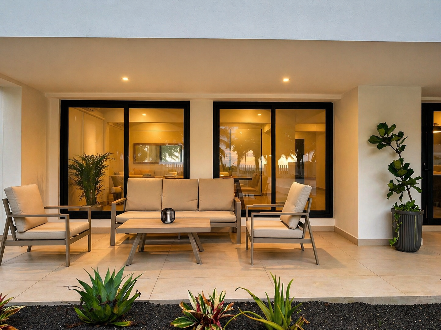 Modern outdoor patio with beige cushioned seating, potted plants, and large glass windows reflecting a sunset and palm trees.