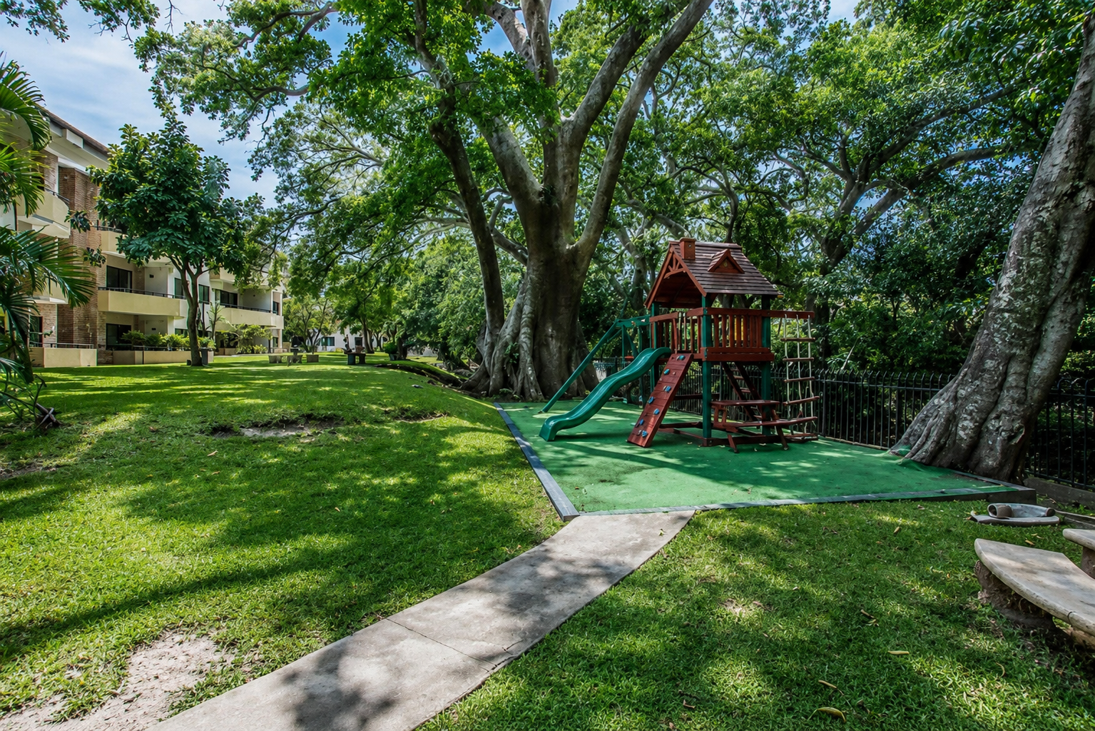 Playground with a slide and climbing structure under large trees in a grassy area, nearby residential buildings.
